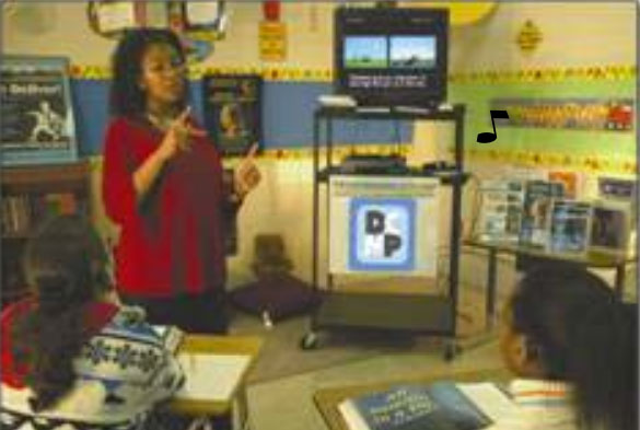 A teacher stands next to a television displaying a captioned video.