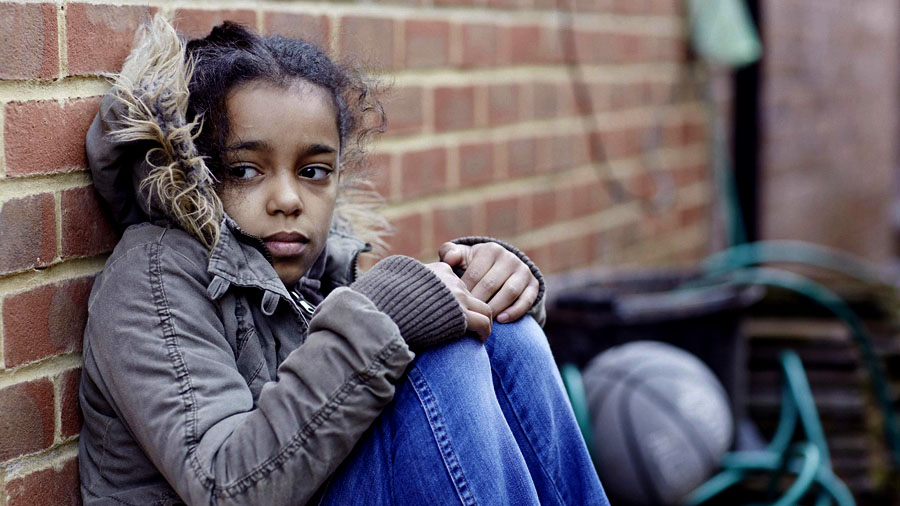 A young black girl wears a winter coat, sitting against a brick wall.