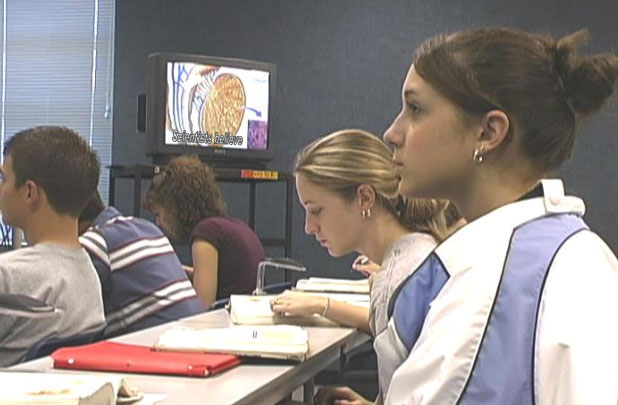 Students sit in a classroom. A TV shows a captioned video.
