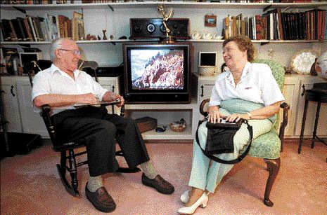 Cody and Margaret Pfanstiehl sit smiling at each other with a television on behind them