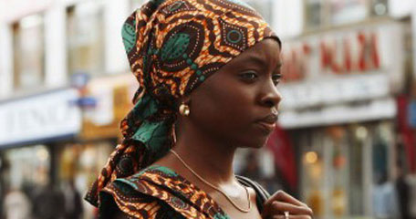 A young black woman wears a head scarf and walks through the city.