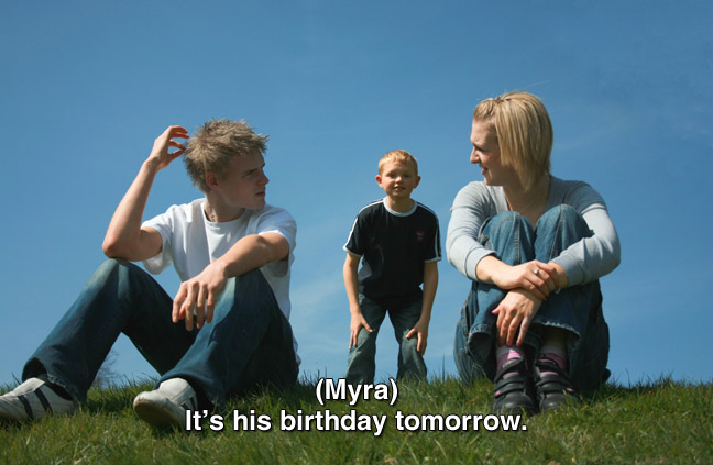 Three teens sitting on grass. caption: (Myra) It's his birthday tomorrow.