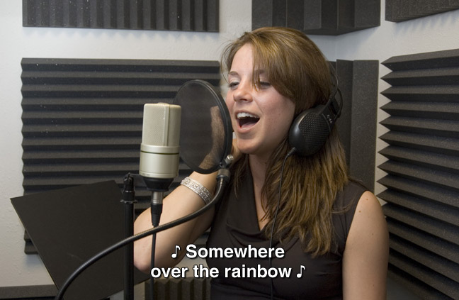 A woman sings in a small studio with a microphone. One musical note is one each end of the captioned sentence. caption: Somewhere over the rainbow.