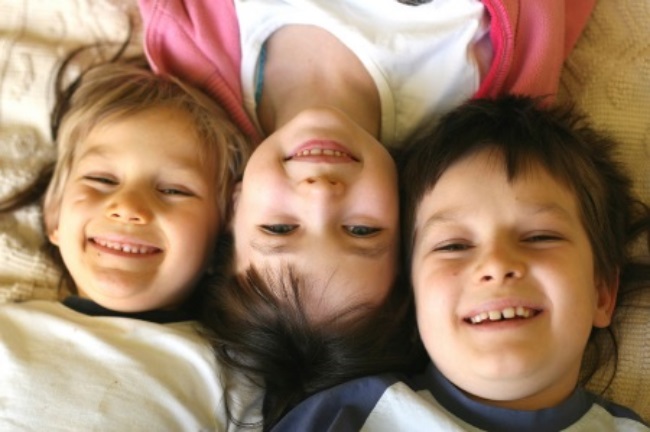 Three young siblings smile lying on the ground, heads together, middle child lying in opposite direction of the other two.