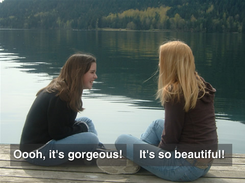 Video still of two girls sitting on a lake dock. Captions read: "Oooh, it's gorgeous!" and "It's so beautiful!".