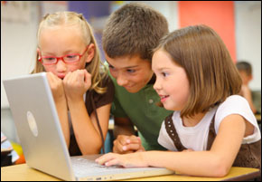 Three grade school children at a desk, smiling excitedly while working on a laptop computer in class.