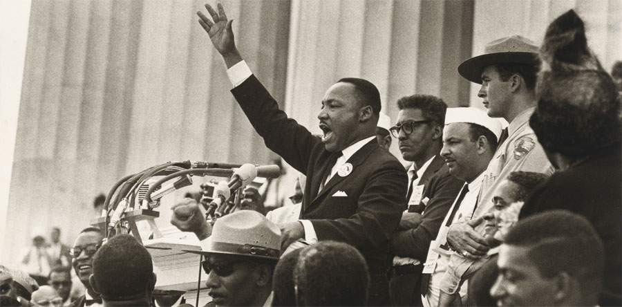 On the steps of the Lincoln Memorial, Martin Luther King, Jr. speaks passionately at a podium filled with microphones, his right arm raised, palm out. He is flanked by several people, including two park rangers.